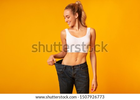 Slim Young Woman In Oversize Jeans Posing Over Yellow Background. Sport And Weight Loss. Studio Shot, Empty Space Royalty-Free Stock Photo #1507926773