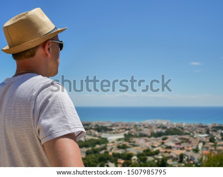 A man in a hat looks in front of him at a beautiful view of a settlement by the sea, close-up