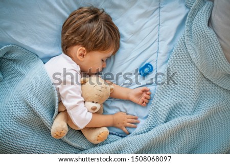 Cute little boy sleeping in bed at home. Sleeping baby in bed, holding a teddy bear. Sweet dreams. Little baby boy sleeping while lying on couch at home. Boy sleeping on bed with teddy bear