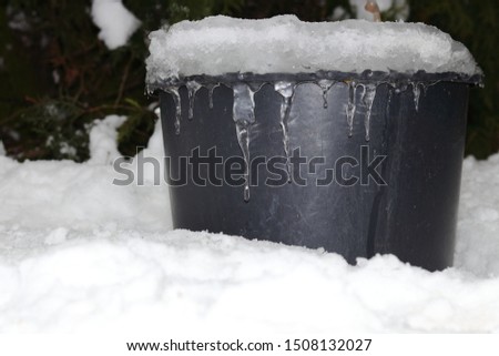Winter atmospheric and motion picture with white snow, ice and mound of icicles on black container (bucket, pail). Plastic receptacle is full of frozen water. Motive of cold, frost and freeze theme.