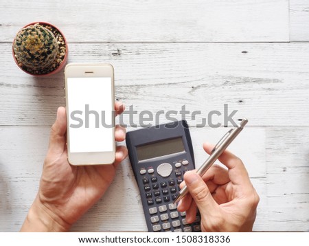 Hand holding smartphone and silver pen with cactus and calculator on white wooden table top view with nature shadow space,Business gadget concept 
