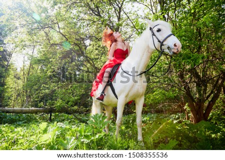 Girl in beautiful red dress on white horse in Park or forest. Photo shoot models and fashion. Unusual posing with an animal in the summer day on nature