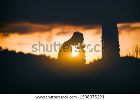 Young man praying and holding Cross under the tree at sunset background. christian silhouette concept.
