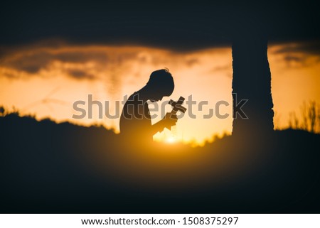 Young man sitting and kneel down for pray and worshipping God with cross under the tree at sunset background. christian silhouette concept.