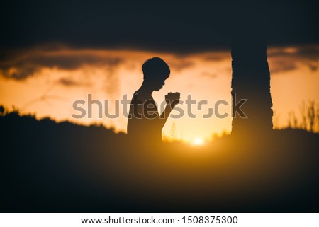 Young christian sitting and kneel down for pray under the tree at sunset background. christian silhouette concept.