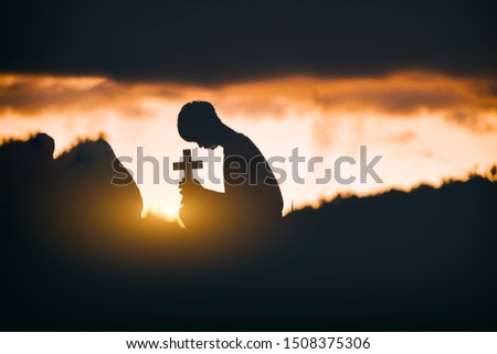 Young man praying and holding Cross In front of the stone at sunset background. christian silhouette concept.