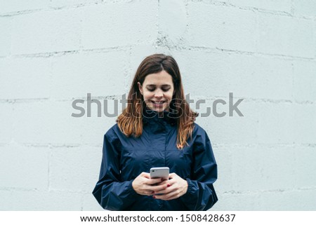 a frontal photo of a young woman using the smartphone in front of a white wall with a blue coat