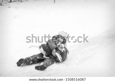 In winter, on the ice, a rustic little boy with a dog in his arms slides down the hill. Black and white photo.