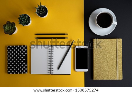 Flat lay office desk of modern design with office supplies. Smartphone with screen and notebook with blank paper and cup of coffee on yellow and balck background. Top view.