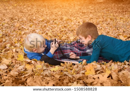 Two little brothers take pictures of each other, lying in yellow autumn leaves. Fall day