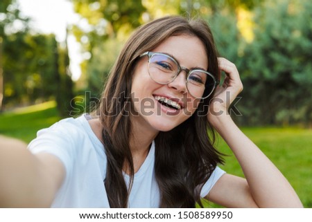 Close up of a smiling pretty girl sitting at the park, taking a selfie