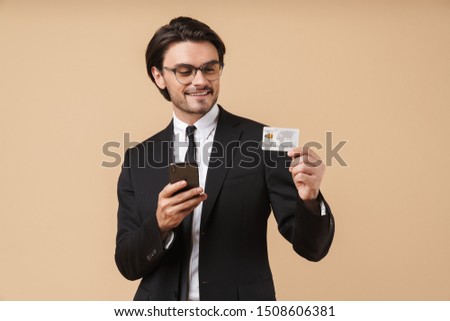 Image of successful businessman in formal suit holding cellphone and credit card isolated over beige background