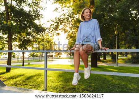 Image of romantic happy woman dressed in casual wear smiling and sitting on railing in green park