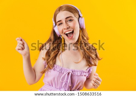 Photo of a young smiling happy girl isolated over yellow wall background listening music with earphones.