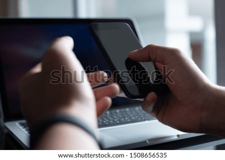 Mock up, man thumb touching on blank screen smart mobile phone, working on laptop computer on office desk as background, use for internet payment mobile application or web design