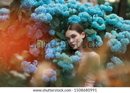 Image of attractive calm girl with brunette hair and bright makeup wearing pink t-shirt, standing among blue flowers and looking at the camera, outdoor photo