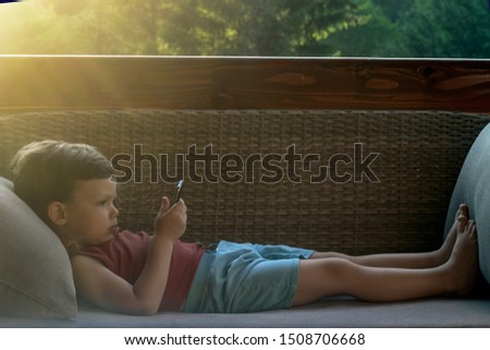 Little cute curious boy with brown hair in light summer clothes,relaxing on couch and watching phone. Beautiful boy holding mobile phone in hands.Toddler playing games, watching cartoons on smartphone