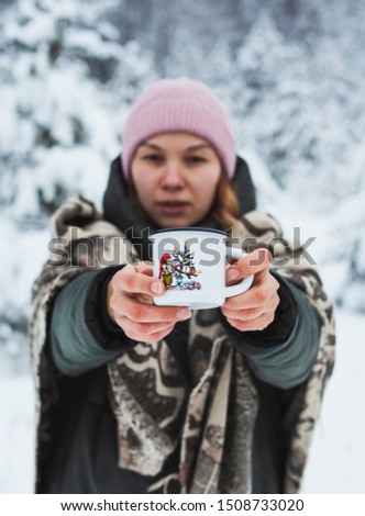 Young woman in pink hat demonstrating metal white cup with funny cartoon on snowy winter forest background. Hipster girl holding hot drink outdoors on nature. Active lifestyle leisure holidays