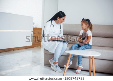 Female doctor with notepad sits in the clinic with little girl.