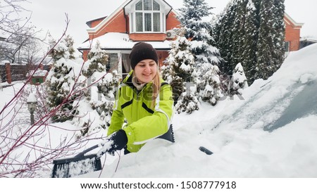 photo of smiling girl in green coat cleaning a snow covered car after blizzard