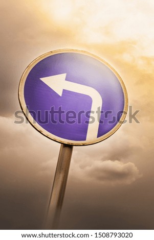 Turn left ahead sign, blue round roadside traffic signage, against the dramatic sky