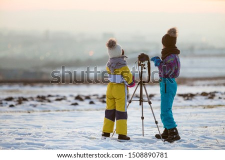Two children boy and girl having fun outside in winter playing with photo camera on a tripod on snow covered field.