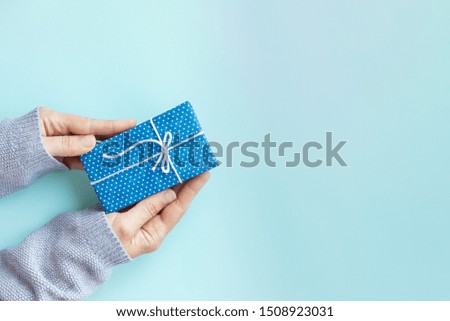 Woman hands in cozy gray sweater holding gift box. Small present in polkadot blue craft paper decorated with bow on blue background. Copy space.