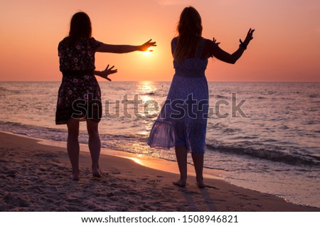 Rear View Of Two Young Women Looking At Sea At Sunset and Dancing. Silhouettes of two young women dancing at beach at colorful sunset