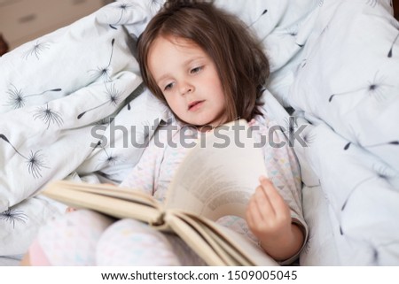 Cute girl reading book siting in her bed and readsinteresting fairy tales, turning pages and finds colourful pictures or interesting stories, charming kid lying on pillows. Childhood concept.