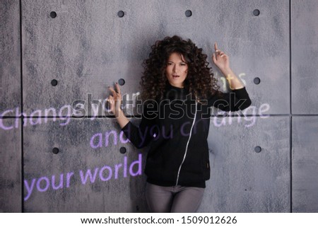 Curly young woman actively posing on a concrete wall background. A colored light sign is projected onto the wall that reads: Change your thoughts and you change your world. Copyspace.