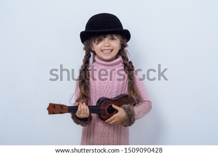 Little girl playing guitar on a grey wall background. Funny kid girl with ukulele guitar. Fashionable country girl playing music. 