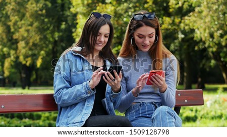 Portrait of two female friends sitting on a bench in park and using mobile phone, choosing pictures for social media app