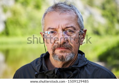 A close-up portrait of an intelligent elderly man with a serious face, glasses with a thin metal frame, a gray beard and a short haircut. Photo of an old man on a blurred green park background. Royalty-Free Stock Photo #1509129485