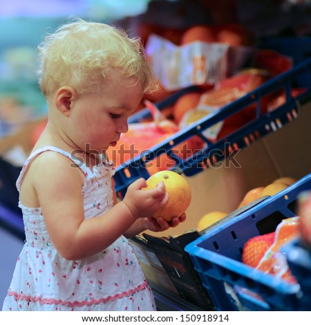 Adorable little baby girl in beautiful dress is helping mother with shopping, she is standing looking at orange that she picked from a shelf in vegetables and fruits department in a supermarket