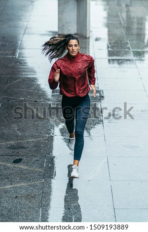 frontal photo of a sportswoman running wearing a red windbreaker, black leggings and white trainers under the rain in the city