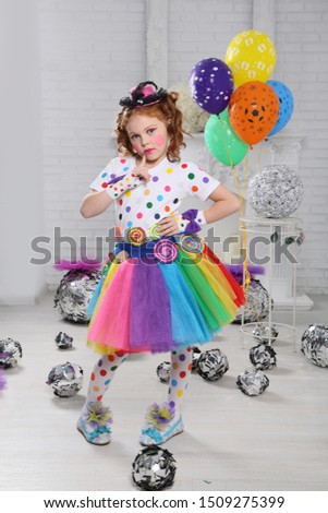 Little clown with balloons. A girl with red hair with bright makeup and colorful clothes plays a clown . The white room . posing standing .