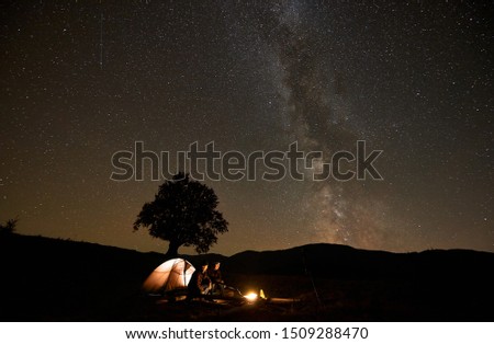 Two tourists hikers men sitting at burning campfire in front of brightly lit from inside tent, photo camera on tripod on green grassy valley under dark blue starry sky with Milky Way constellation.