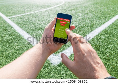 A man's hand holding a mobile phone in which the interface of a sports betting page is seen, on the green background of a soccer field. Online betting concept.
