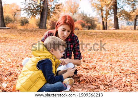 Small boy and his mother relaxing in autumn leaves and using smart phone at the park.