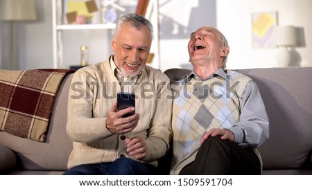 Elderly man showing funny photo on smartphone aged friend, having fun at home