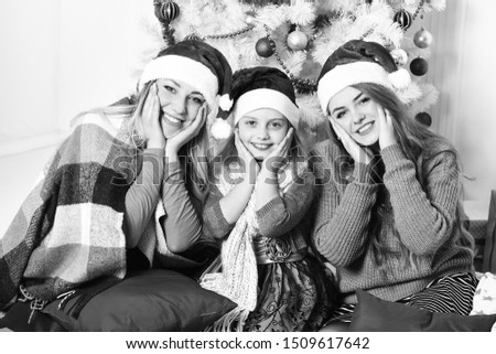 Family or friends in Santa hats near present boxes. Girls with happy smiling faces with white and blue Christmas tree on background. Winter holiday and party concept. Sisters sit holding cheeks