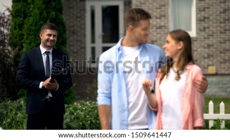 Estate broker watching happy couple enjoying new house showing keys to camera