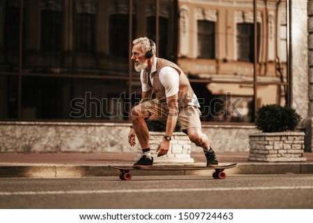 Portrait of bearded hipster man in white shirt on city street. Stylish happy model ride on longboard near road on buildings background Royalty-Free Stock Photo #1509724463