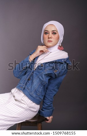 Fashionable young woman in white pants, long sleeves jeans jacket and hijab isolated on grey background. Stylish Muslim female hijab fashion lifestyle portraiture concept.