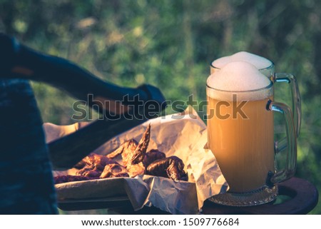 Glass of light beer against background of green park trees outdoor picnic, outing party rest on nature. Snack sausage, smoked chicken wings, vintage toning picture