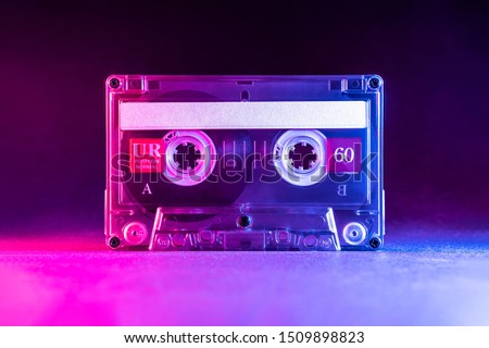 Transparent audio cassette tape lit by pink and blue lamps on a black background. Front, top view. Royalty-Free Stock Photo #1509898823