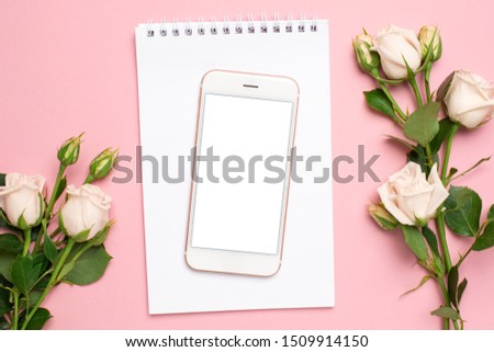 Mobile phone with a white notebook and roses flowers on pink background
