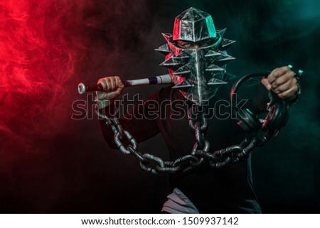 Mysterious man in a helmet with a bat and chains in black wear. Fantasy book or computer game cover concept on halloween disco party.