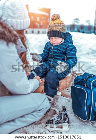 little boy of 3.5 years old, winter on city skating rink, sits on bench, woman s mother changes her child s clothes, puts on shoes, rest on weekend, snow drifts, warm clothes, hats, jackets