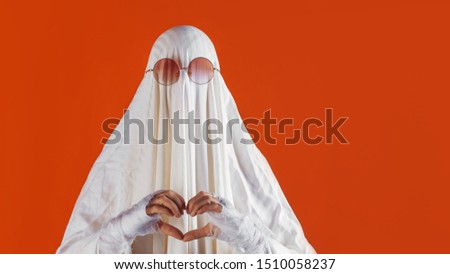 Happy Halloween. Cute funny Ghost on a bright orange background. Sheet Ghost costume, Halloween party carnival. Ghost loves you, heart-shaped hand gesture